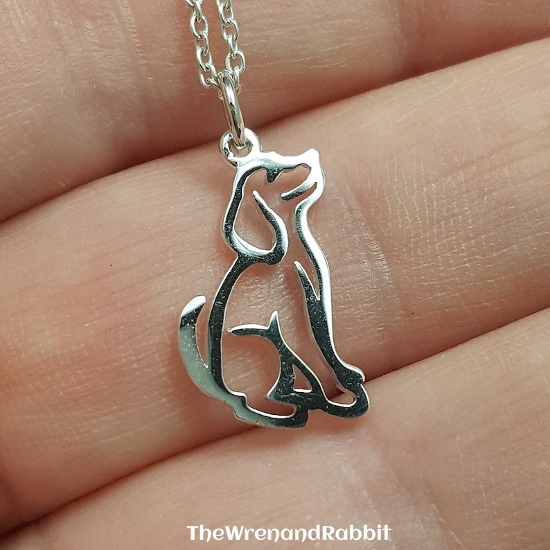 May include: A silver necklace with a cutout design of a dog sitting. The necklace is on a hand.