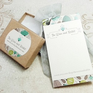 May include: A small brown box with a white circle sticker that says "The Wren and Rabbit" with a green and blue floral design. The box is sitting on a white surface. A white card with a green and blue floral design and the text "The Wren and Rabbit" and "www.etsy.com/shop/thewrenandrabbit" is sitting on a white surface.
