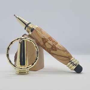Puede incluir: Un bolígrafo de madera con un diseño de Estrella de David y detalles dorados. El bolígrafo tiene una punta dorada y un extremo de lápiz táctil negro. También se incluye un llavero dorado.