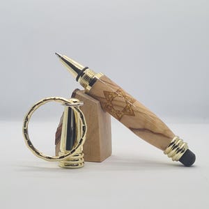 Puede incluir: Un bolígrafo de madera con detalles dorados y un diseño de Estrella de David. El bolígrafo descansa sobre un soporte de madera junto a un portalápices dorado. El bolígrafo tiene una punta de lápiz táctil negra.