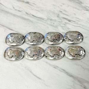 May include: Eight silver-toned oval-shaped buttons with a hammered texture. Each button features a raised sheep design in the center. The buttons are arranged in two rows of four on a white marble surface.