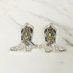 May include: Two silver-toned cowboy boot charms with clear rhinestones and colorful enamel details. The boots have a decorative design on the shaft with yellow, green, and blue accents. The charms are suitable for jewelry making.