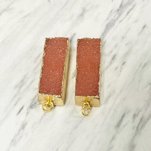 May include: Two rectangular, reddish-pink gemstone pendants with gold-colored edges and loops. The pendants have a textured, crystalline appearance and are set against a white and gray marble background.