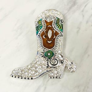 May include: A silver-toned cowboy boot brooch, encrusted with clear rhinestones. The boot features brown, green, and white enamel detailing. A spur with a rhinestone accent is attached to the heel. A decorative accessory.
