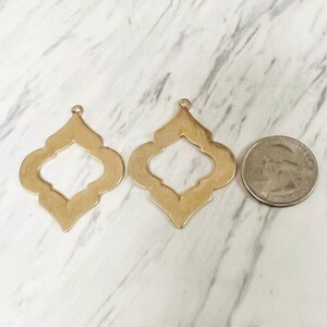 Op de afbeelding: Twee goudkleurige, openwerk, Marokkaanse stijl hangerbedels met een munt voor maatvergelijking. De bedels hebben een vierpasvorm met een kleine lus aan de bovenkant om op te hangen. De achtergrond is een wit en grijs marmerpatroon.