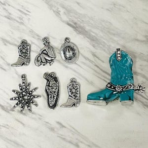 Op de afbeelding: Een verzameling zilverkleurige cowboy-thema bedels en een turquoise cowboylaars. Bedels zijn onder meer een cowboy, een cowboyhoed, een spoor en twee cowboylaarzen. De turquoise laars heeft een zwart bloemmotief.