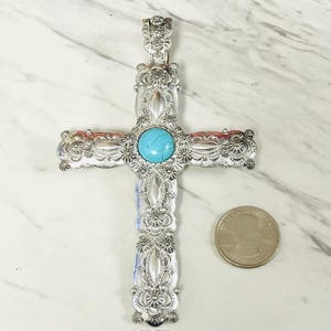 May include: Silver-toned cross pendant with intricate floral and scrollwork details. A round, turquoise-colored stone is set in the center. The cross is shown next to a quarter for size comparison.