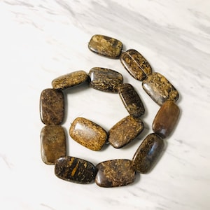 Puede incluir: Una hebra de cuentas rectangulares de piedra preciosa de color marrón y dorado. Las cuentas están hechas de una piedra natural con un patrón moteado.