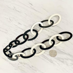 May include: A decorative chain composed of alternating oval links in solid black and off-white colors. The chain is arranged in a curved line next to a silver-colored United States quarter coin for scale, showing the relative size of the individual links.