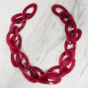 May include: A chunky necklace composed of large, interlocking oval-shaped links in a glossy, deep magenta red color. The chain features a smooth, reflective finish and is arranged in a circular formation with an open end at the top.