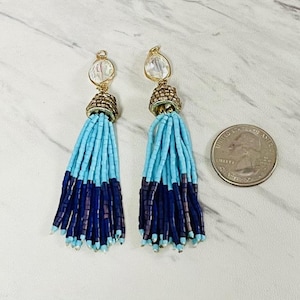 May include: A pair of dangle earrings featuring clear faceted stones at the top, gold-toned metal caps, and long tassels made of light blue and dark blue seed beads. A United States quarter is placed next to the earrings for size reference.