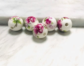 May include: Five white ceramic beads with a floral design. The beads have a hole in the center and are decorated with pink and purple flowers and green leaves.