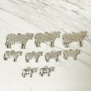 May include: A collection of silver-colored cow charms. The charms are in various sizes and have a rustic, textured finish. Each charm has a small loop at the top for attaching to a chain or other craft project. The cows are in a standing position.