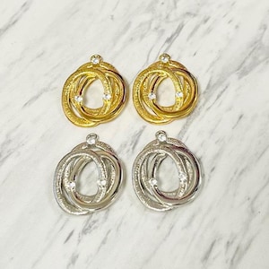 May include: Four pairs of earrings in gold and silver tones. Each earring features an intertwined circular design with small, clear gemstones. The earrings are displayed on a white and gray marble surface, showcasing their intricate details and elegant style.