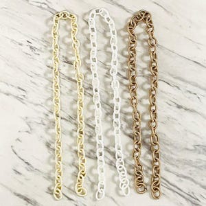 May include: Three long, flexible chain straps arranged vertically. The left strap is a light cream color, the center strap is solid white, and the right strap is a metallic bronze tone. Each strap consists of repeating oval-shaped links.