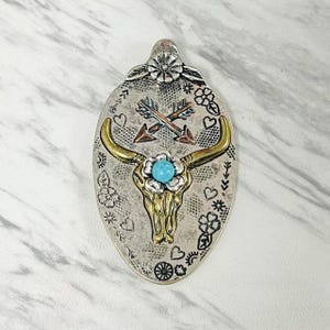 以下が含まれることがあります： 金色の雄牛の頭蓋骨、ターコイズブルーの中央、交差した矢が特徴の銀色のペンダント。ペンダントには花とハートのアクセントがあり、上部に花があります。ペンダントは楕円形です。