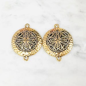 May include: Two circular gold-toned metal connectors featuring a hammered outer edge and a central openwork filigree design. Each piece includes a small loop at the top and bottom for attachment, showcasing a textured finish and intricate metalwork.