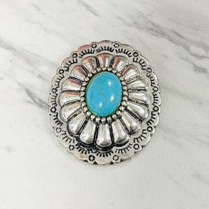 May include: Oval-shaped silver-toned metal button with a turquoise-colored stone center. The button has a detailed, floral-like design around the stone. The button is likely used for clothing or accessories.