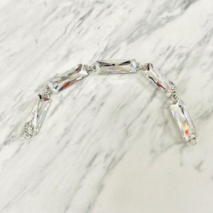 May include: A silver bracelet featuring rectangular, clear crystal stones. The crystals are linked together with small silver connectors. The bracelet is displayed on a white and gray marble surface, showcasing its reflective qualities.