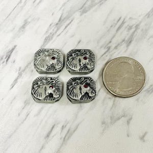 May include: Four square silver-coloured metal beads, each featuring a relief design of a horse head with a small red stone eye. The beads are arranged in a two-by-two grid next to a United States quarter coin for scale, showing their relative size.