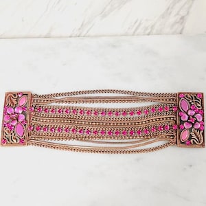 May include: A wide cuff bracelet featuring a copper-toned metal finish with multiple layered chains and rows of vibrant pink rhinestones. The bracelet is anchored by two rectangular end pieces decorated with floral motifs and pink cabochon stones.