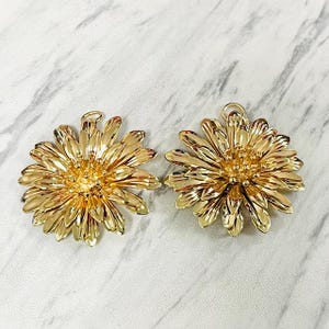 May include: Two gold-colored daisy-shaped pendants. Each pendant features detailed petals radiating from a textured center. The pendants have a loop at the top for hanging. The background is a light marble pattern.