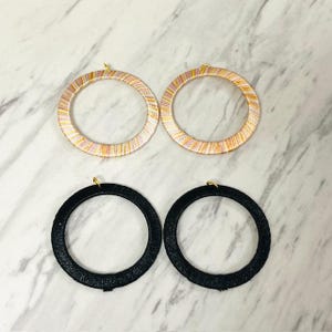 May include: Four hoop earrings. Two are black, textured, and circular. The other two are circular with a woven pattern of pink, yellow, and white stripes. Each earring has a small gold-colored hook.