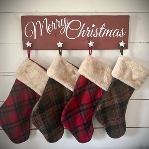Könnte beinhalten: Vier rote und grüne karierte Weihnachtsstrümpfe mit weißem Kunstfellbesatz hängen an einem Holzschild mit der Aufschrift "Merry Christmas".