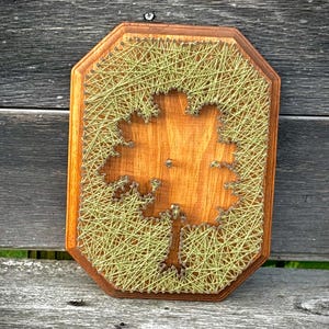 May include: Wooden string art of a tree on a brown, octagonal wooden plaque. The tree is cut out of the wood, and the outline is filled with green string. The plaque is mounted on a weathered wooden surface.