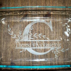 Puede incluir: Un plato para hornear de vidrio transparente con un borde azul. El plato está grabado con la letra "C" rodeada de diseños florales y el texto "Theresa's Kickasserole".