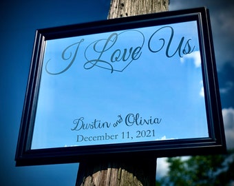 May include: A framed sign with the words "I Love Us" in elegant script, with a heart symbol. Below, it reads "Dustin and Olivia, December 11, 2021." The background is a blue sky with clouds.