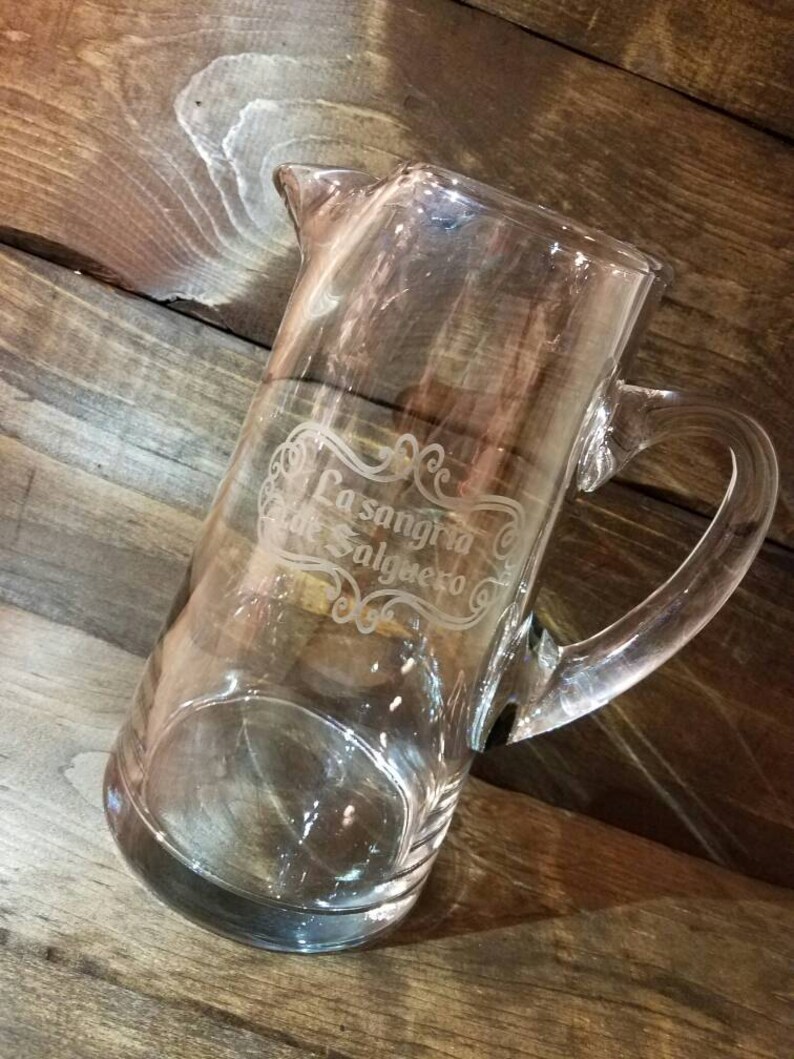 May include: A clear glass pitcher with a handle, etched with the text "La Sangria de Salgueiro".