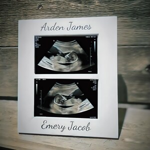 May include: White picture frame with two black and white ultrasound images. The top image has the name "Arden James" written above it. The bottom image has the name "Emery Jacob" written below it.