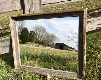 May include: A rectangular, rustic wooden picture frame with a natural, weathered finish. The frame reflects a scenic outdoor view of a grassy field, trees, and a barn under a cloudy sky. The frame is resting on grass.