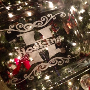 Puede incluir: Un plato para hornear de vidrio transparente con un diseño grabado decorativo que presenta la letra H y el nombre Hoskins. El plato está sentado en un árbol de Navidad con adornos rojos y verdes.