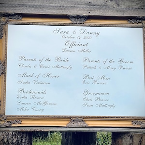 May include: A gold-framed mirror with a white background and black text listing the names of the wedding party. The text includes the names of the bride and groom, the officiant, the parents of the bride and groom, the maid of honor, the best man, the bridesmaids, and the groomsmen. The date of the wedding is also listed: October 14, 2022.