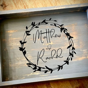 May include: A gray wooden serving tray with black handles. The tray is decorated with a black wreath design and the names "Matthew and Rachel" in a cursive font.