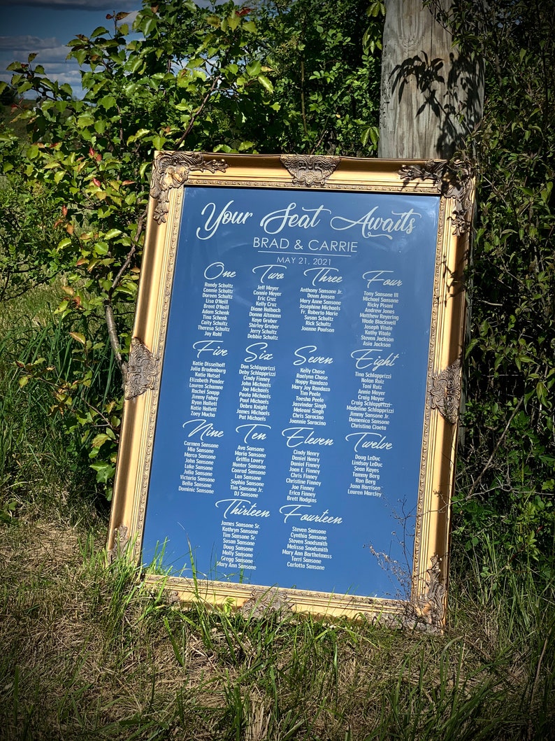 May include: A gold-framed mirror with a blue background lists the names of wedding guests for a wedding reception seating chart. The text at the top reads "Your Seat Awaits Brad & Carrie May 21, 2021".