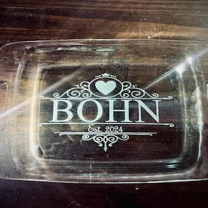 Puede incluir: Un plato para hornear de vidrio transparente con un diseño blanco que dice "BOHN Est. 2024" con un corazón sobre el nombre.