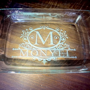 Puede incluir: Un plato para hornear de vidrio transparente con asas, que presenta un diseño grabado de un monograma "M" con el nombre "MONYEI" debajo.