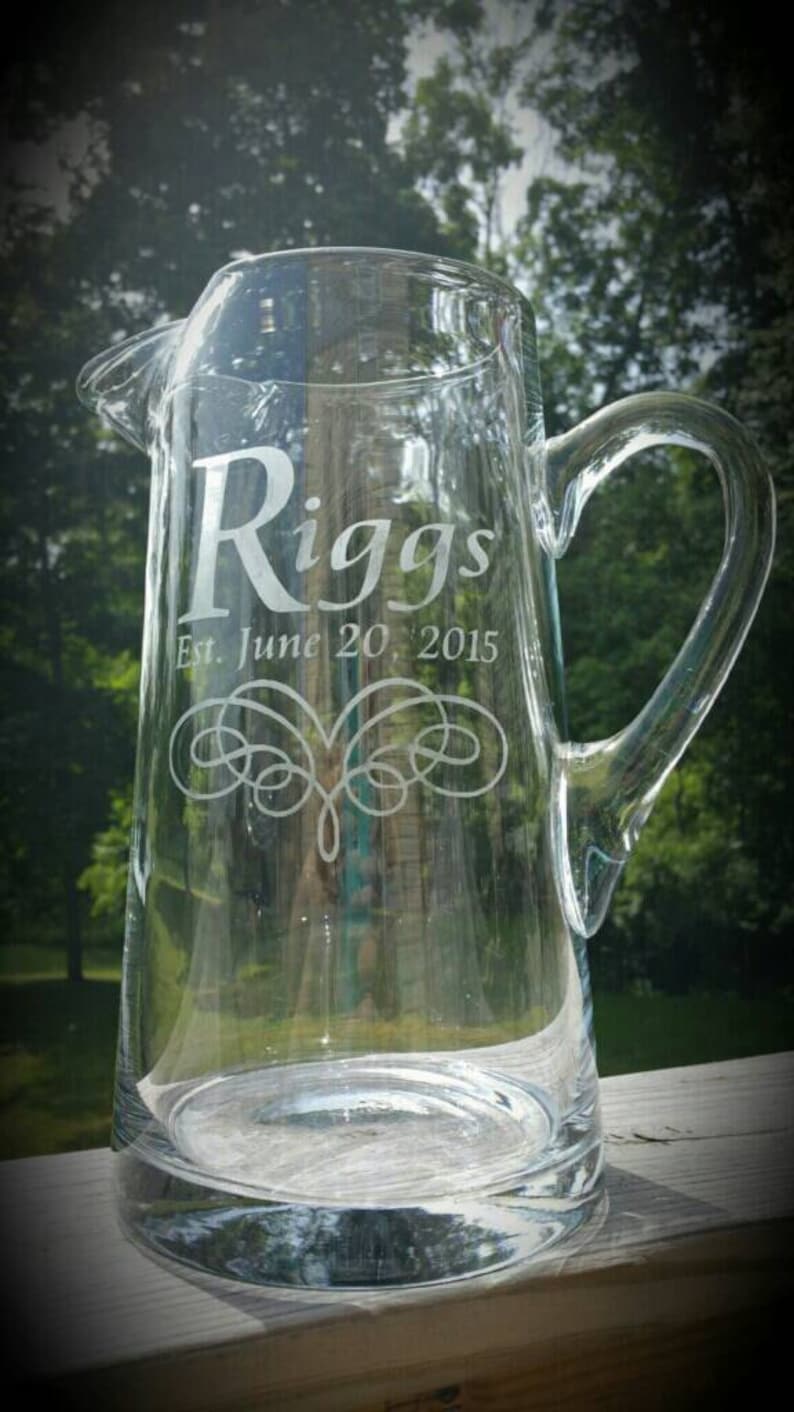 May include: A clear glass pitcher with a handle, engraved with the text "Riggs Est. June 20, 2015" and a decorative swirl design.
