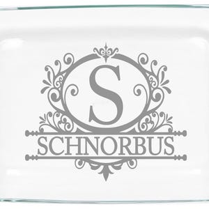Puede incluir: Una fuente para hornear de vidrio transparente con esquinas redondeadas y asas. El centro presenta un diseño decorativo gris con la letra "S" dentro de un marco ovalado y el nombre "SCHNORBUS" debajo.