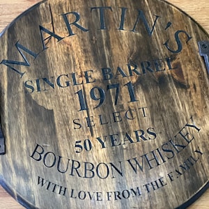 Può includere: Un vassoio rotondo in legno con macchia marrone scuro e scritte nere. Il testo recita "MARTIN'S SINGLE BARREL 1971 SELECT 50 YEARS BOURBON WHISKEY WITH LOVE FROM THE FAMILY." Presenta due maniglie in metallo.