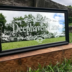 Może przedstawiać: Czarne lustro w ramie z białym grawerowanym wzorem i napisem "Deephaven Lane" na szkle.