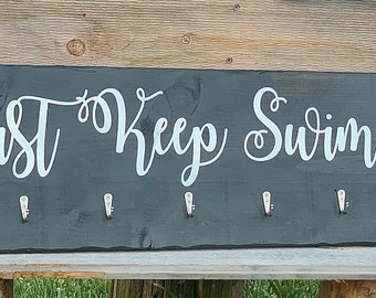 Könnte beinhalten: Ein dunkelgraues Holzschild mit dem Text "Just Keep Swimming" in weißer Kursivschrift. Das Schild hat acht Metallhaken zum Aufhängen von Gegenständen.