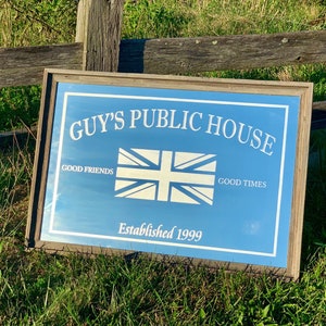 Könnte beinhalten: Ein blaues und weißes Schild mit dem Text "GUY'S PUBLIC HOUSE" in weißen Buchstaben. Das Schild zeigt auch eine weiße Union Jack Flagge und den Text "GOOD FRIENDS" und "GOOD TIMES" auf beiden Seiten der Flagge. Das Schild enthält auch den Text "Established 1999".