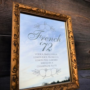 May include: A gold framed mirror with a white inscription that reads "French 72" and a list of ingredients for a cocktail. The inscription includes "Vodka, Simple Syrup, Lemon Juice, Prosecco, Lemon Peel Garnish, Served in a Champagne Flute".