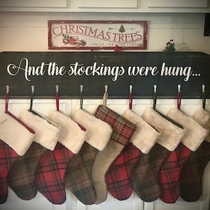 Puede incluir: Un letrero de madera negro con el texto "And the stockings were hung..." colgado sobre una fila de ocho calcetines de cuadros rojos y verdes con ribete de piel blanca.