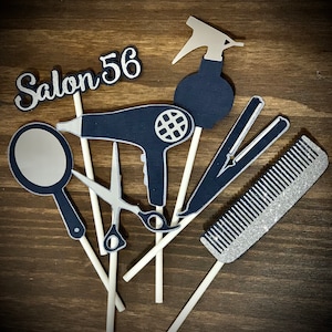 May include: A set of six cake toppers for a salon themed party. The toppers are made of paper and feature a silver comb, a silver hair dryer, a silver pair of scissors, a silver hair straightener, a silver spray bottle, and a silver sign that says "Salon 56".