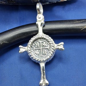 May include: A silver-toned pendant shaped like a cross with a central circular medallion featuring a cross design and textured rope-like border. The arms of the cross resemble elongated bones, and a polished bail is attached to the top.